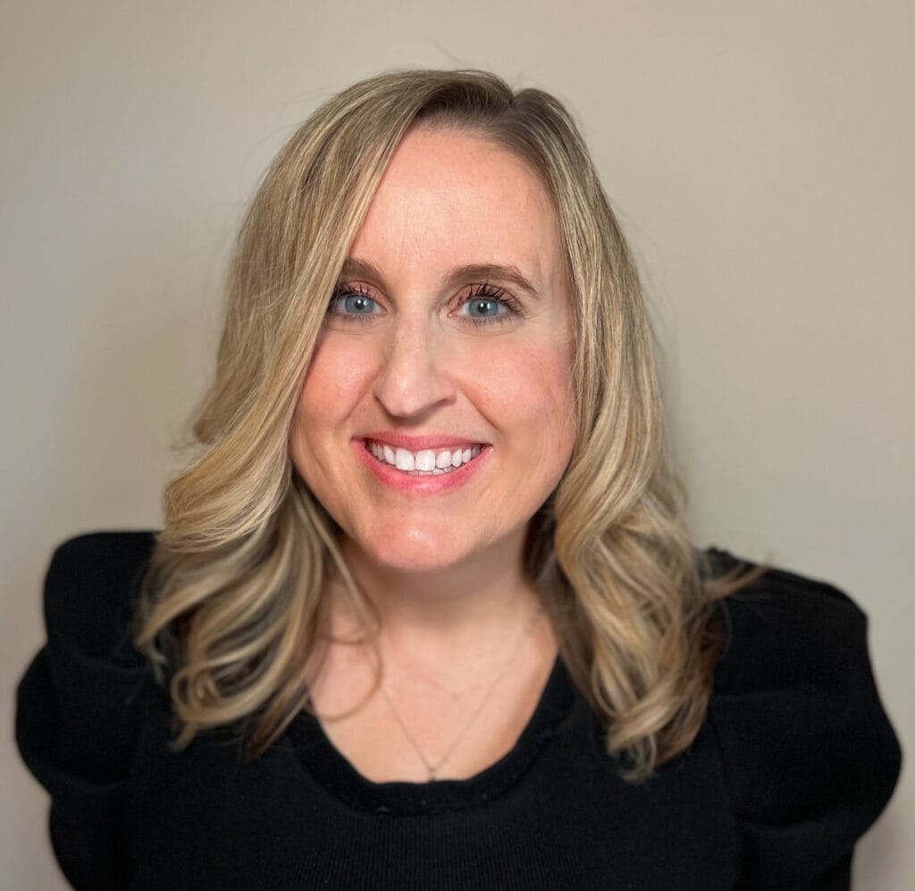 Picture of white woman with shoulder length wavy hair and blue eyes. She is smiling and wearing a black shirt with puffer sleeves.