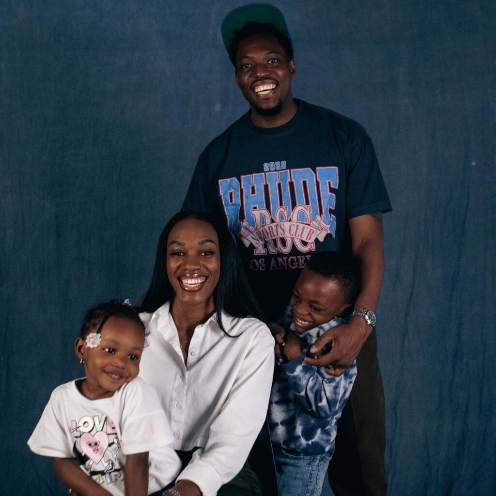 Family photo of a black father smiling, wearing a hat and tee shirt, a black mother in a white button down smiling, and their two kids, boy and girl smiling next to them